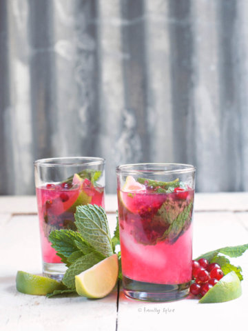 Side view of two glasses of red currant mojito with fresh mint, red currants and lime wedges next to it