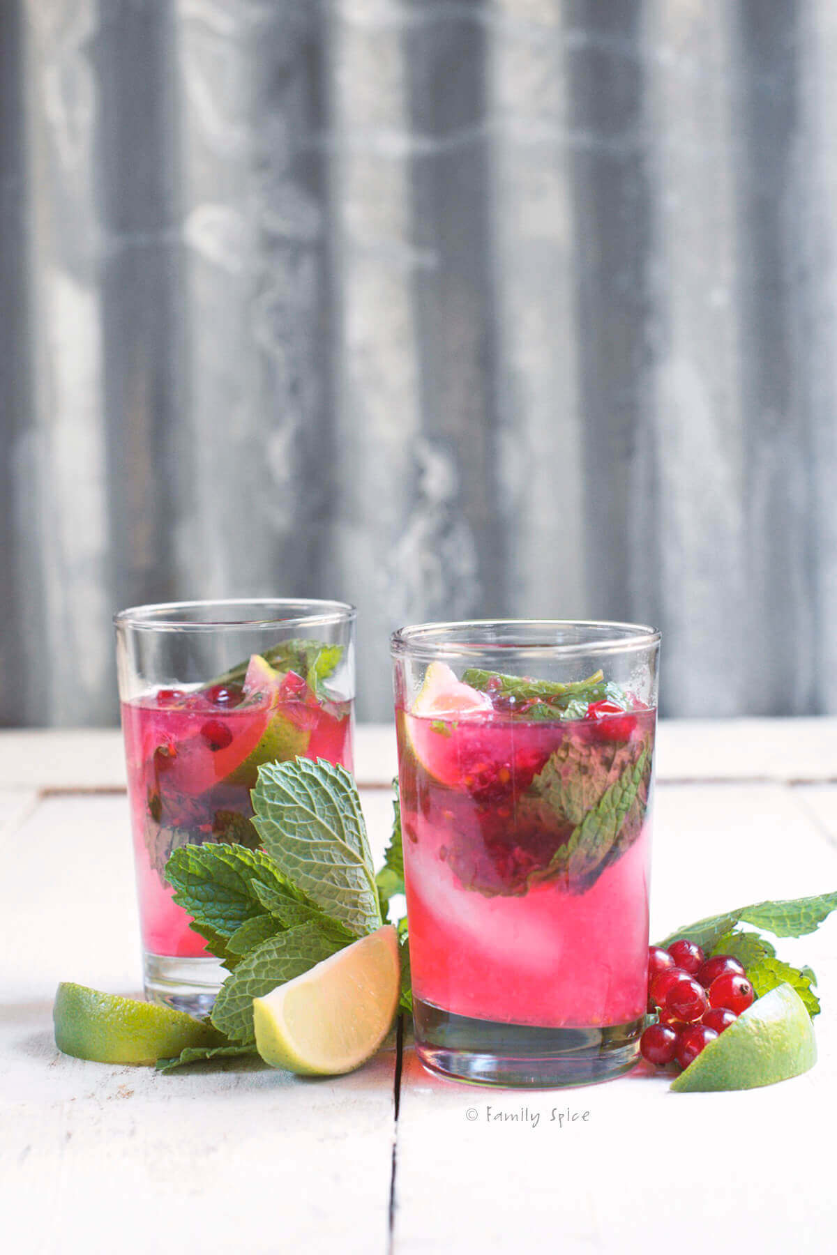 Side view of two glasses of red currant mojito with fresh mint, red currants and lime wedges next to it