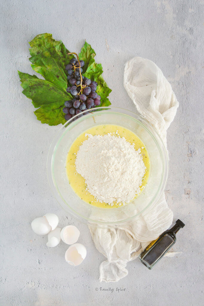 Flour added to cake batter in a glass mixing bowl with egg shells, a bunch of grapes and small bottle of balsamic vinegar next to it