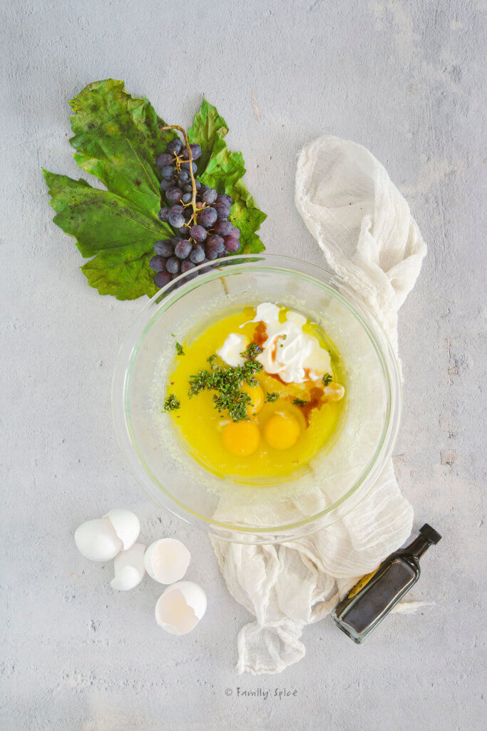 Eggs, yogurt and rosemary added to cake batter in a glass mixing bowl with eggs, a bunch of grapes and small bottle of balsamic vinegar next to it