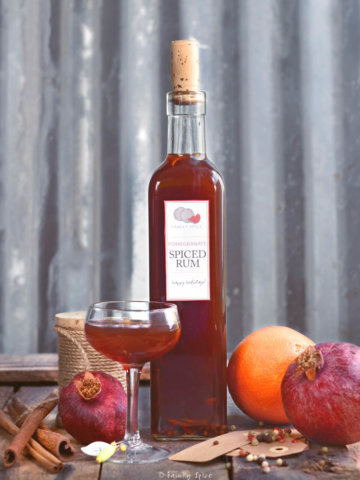 A tall glass bottle with a small stemmed glass with homemade pomegranate spiced rum with ingredients around it