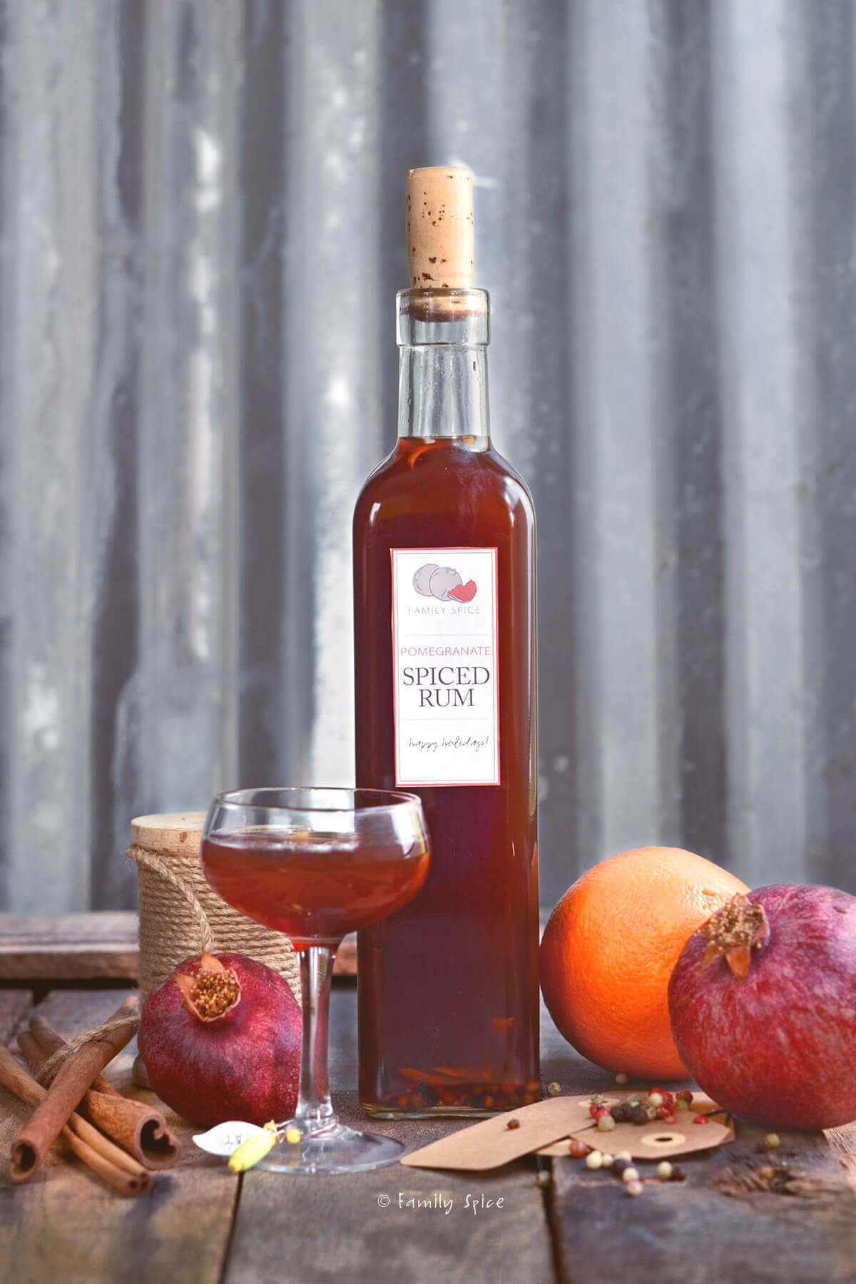 A tall glass bottle with a small stemmed glass with homemade pomegranate spiced rum with ingredients around it