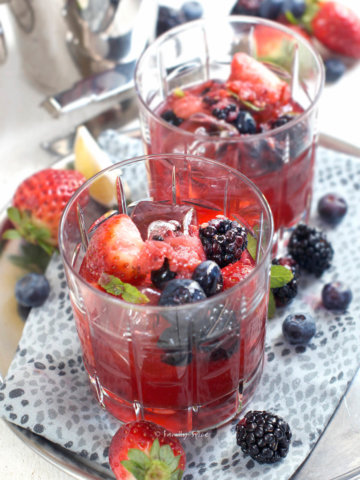 Closeup of a metal tray with two glasses filled with berry bourbon smash with berries around it