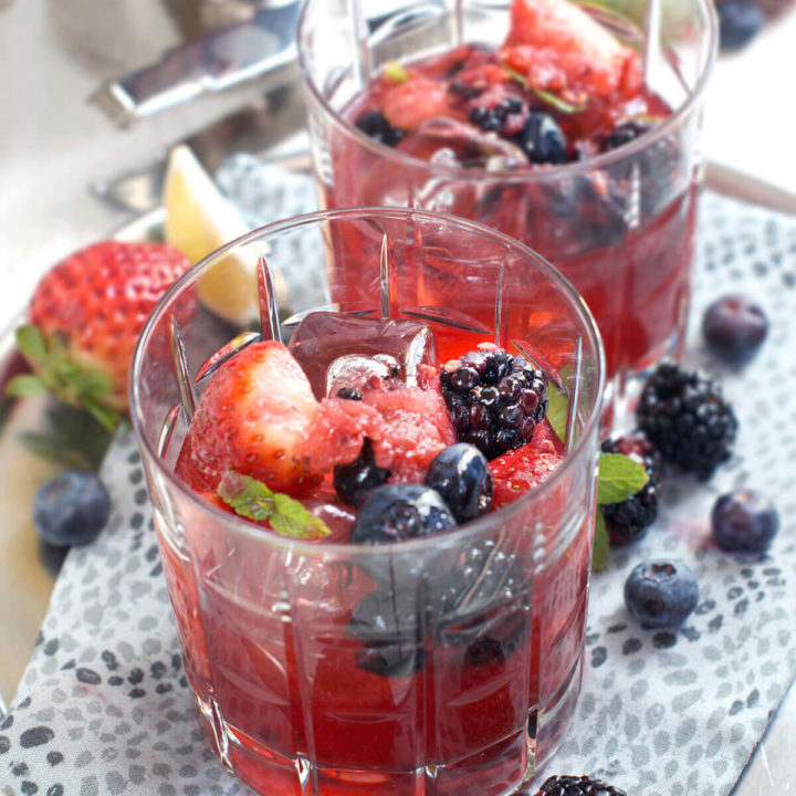 Closeup of a metal tray with two glasses filled with berry bourbon smash with berries around it