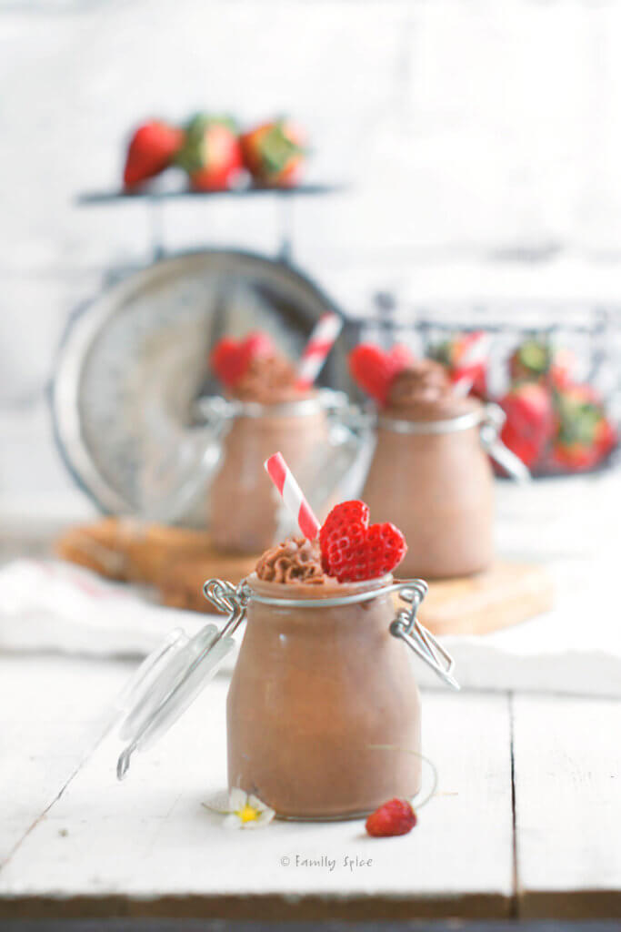 A closeup of a small mason jar filled with chocolate mousse and topped with a strawberry cut in the shape of a heart with more bottles in the background