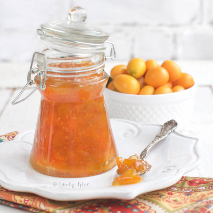 A triangular glass canning jar with kumquat marmalade in it with a bowl of kumquats behind it