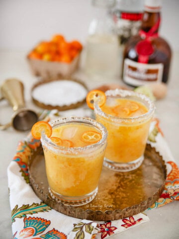 Closeup of two glasses filled with kumquat margaritas on a gold tray with ingredients behind it.