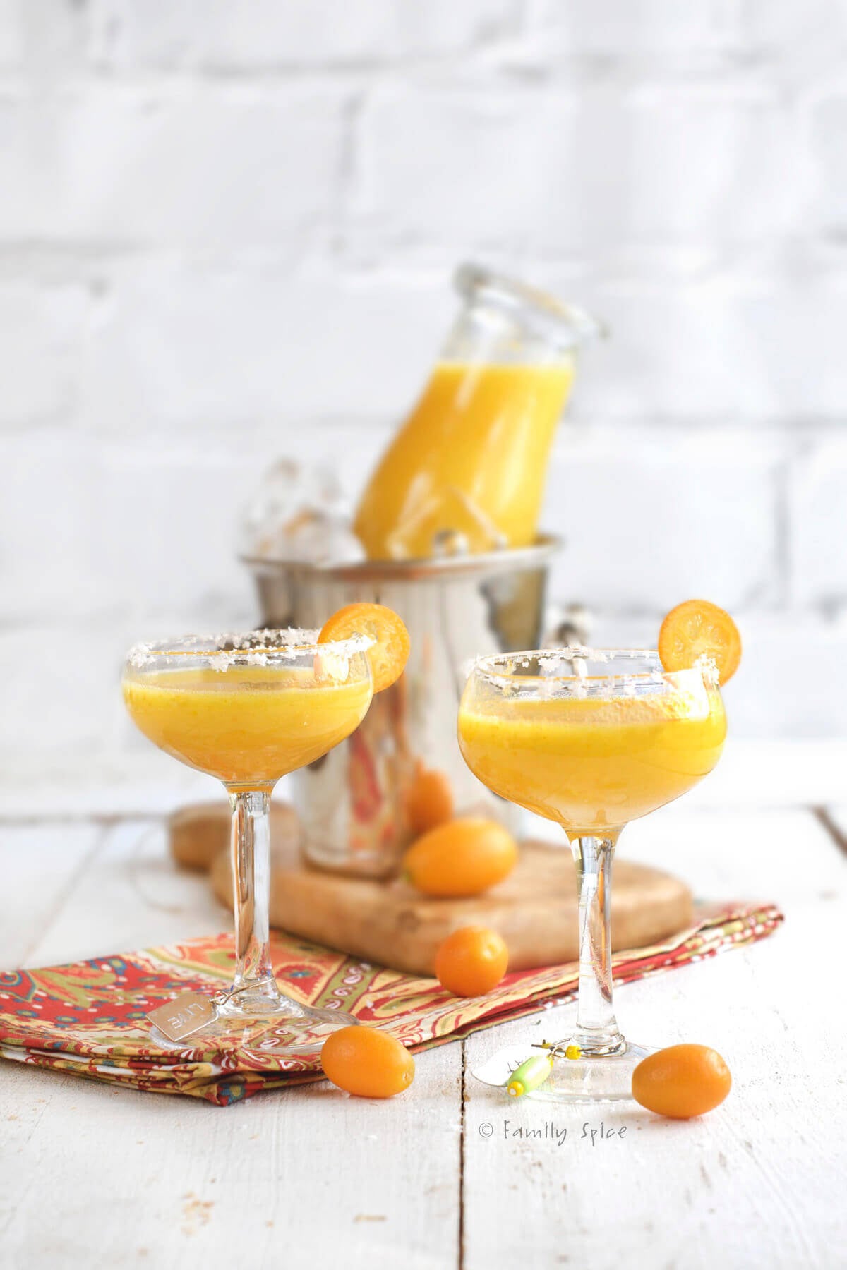 Two small stemwares with kumquat margarita with a small metal ice bucket behind it with a glass bottle of more kumquat margarita