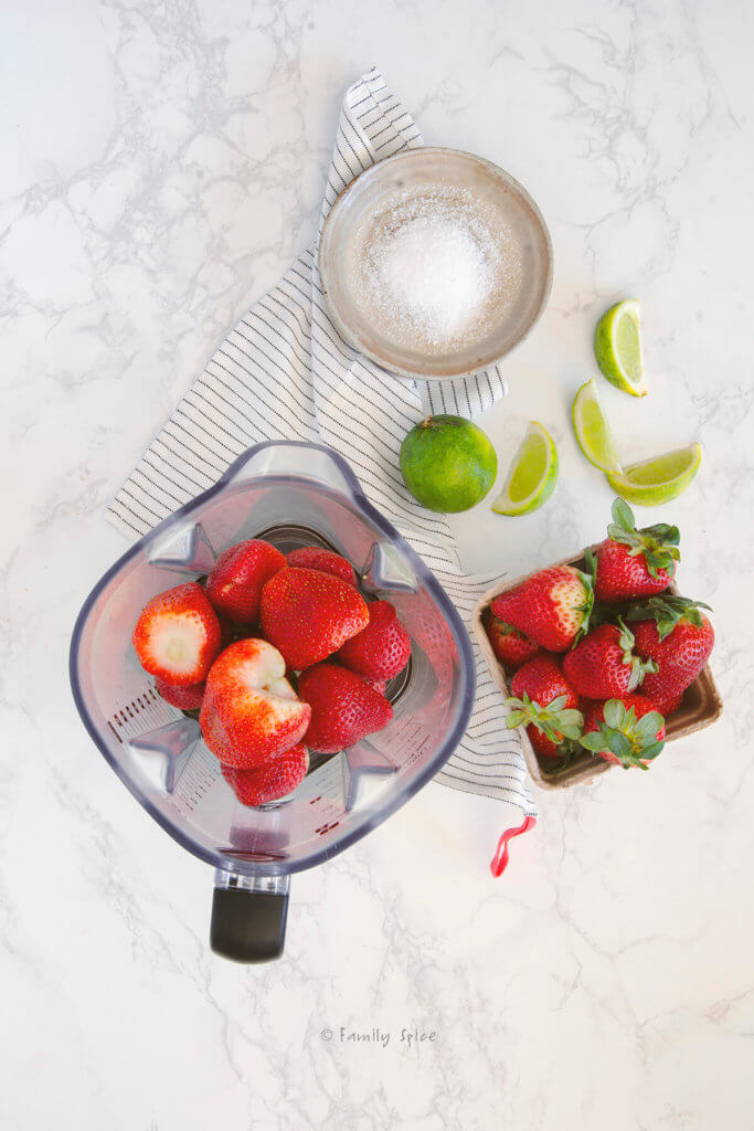 Strawberries in a blender with strawberries, lime wedges and salt next to it