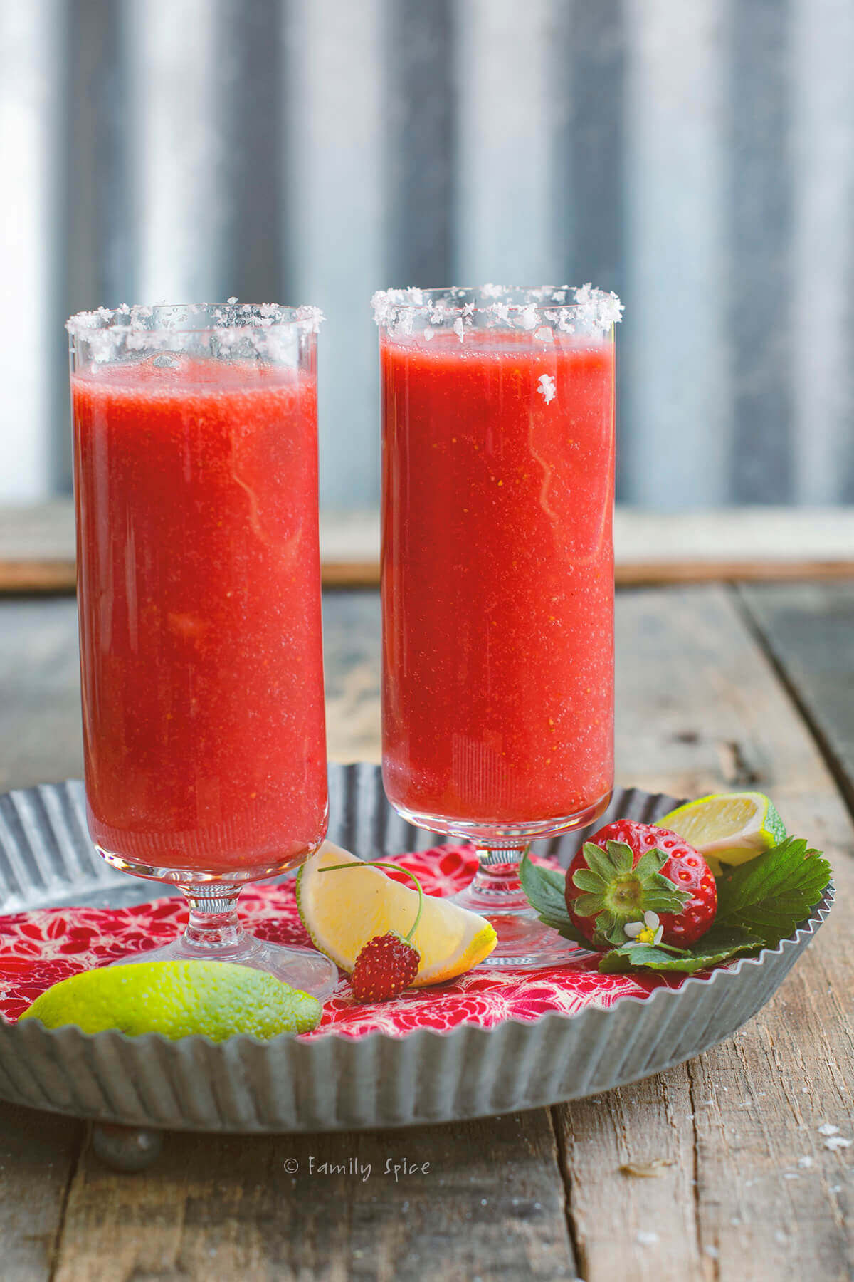 Closeup of two tall glasses with salted rims with salty dog cocktail on a tray with strawberries and limes next to it