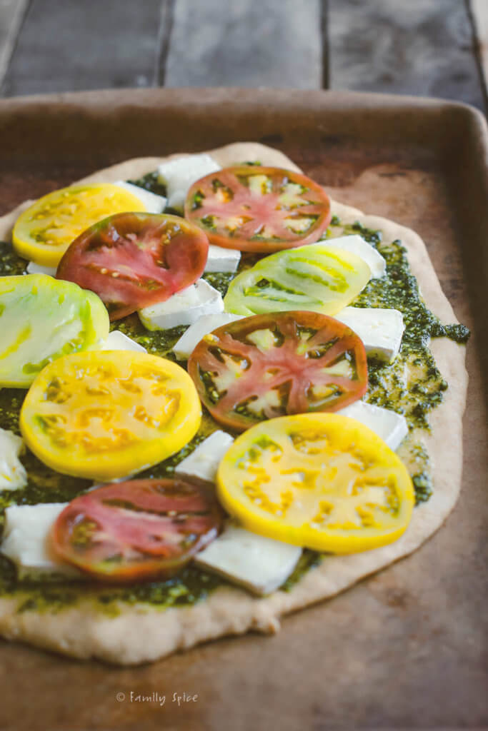 Whole wheat pizza dough on a baking stone with pesto spread on it topped with slices of brie and heirloom tomatoes on top of it
