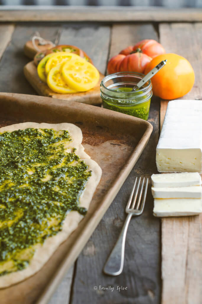 Close up of whole wheat pizza dough on a baking stone with pesto spread on it with a brick of brie, heirloom tomatoes and a jar of pesto next to it