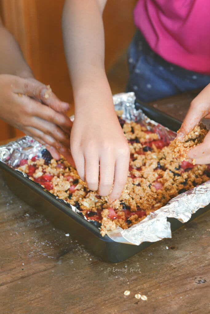 Young girls' hands sprinkling oat crumble over a square baking sheet of whole wheat berry bars ready to bake
