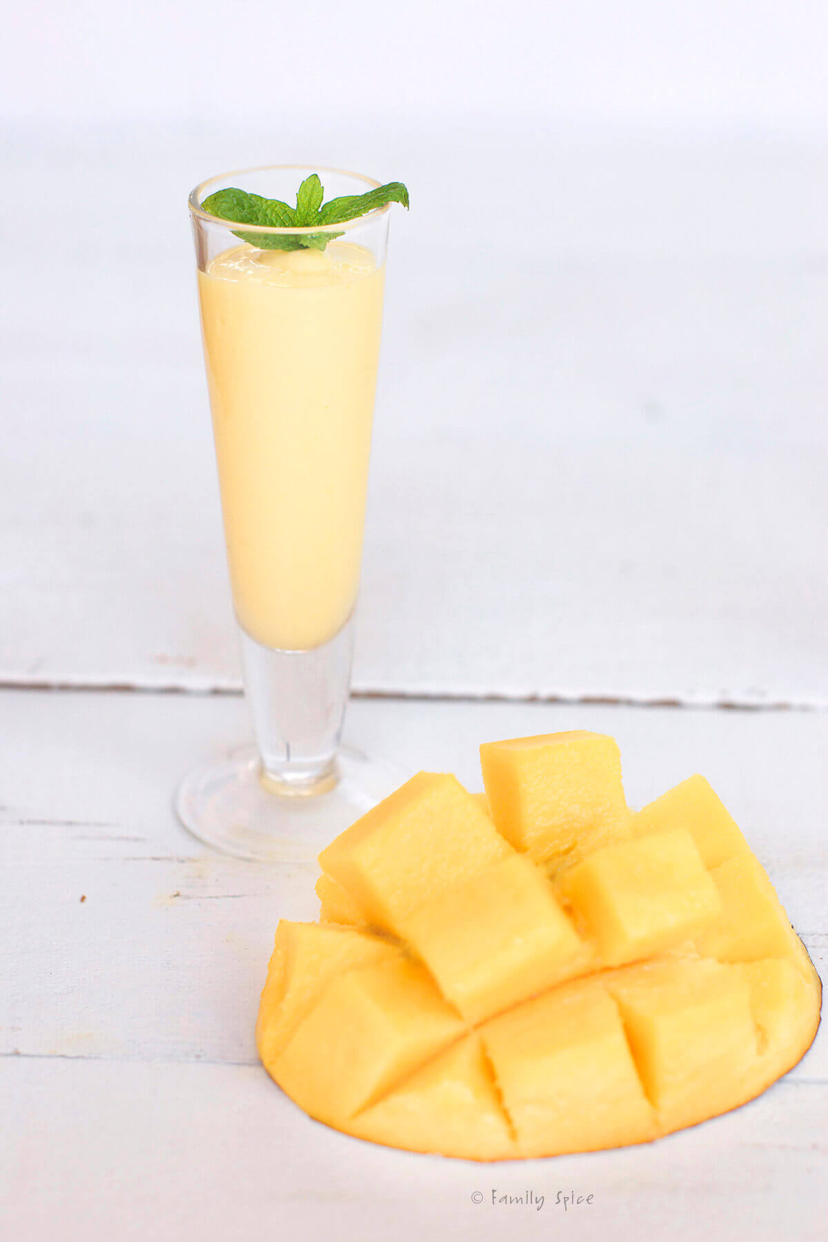 Closeup of a small shot glasses with mango rum smoothie in them and topped with fresh mint next to a halved mango cut into cubes