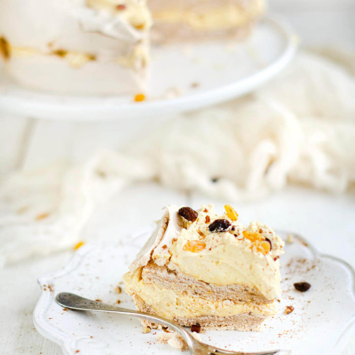 Closeup of a slice of pavlova cake with pumpkin whipped cream between the two layers and on top, garnished with chopped pecans and red and gold raisins