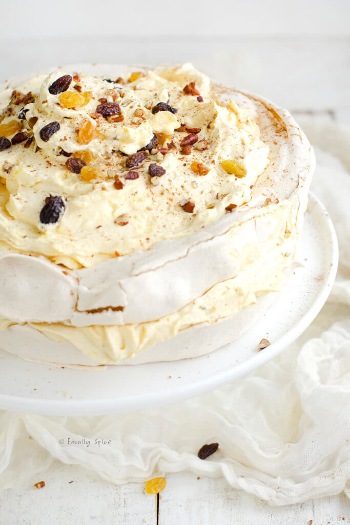 Closeup of a pavlova cake with pumpkin whipped cream between the two layers and on top, garnished with chopped pecans and red and gold raisins