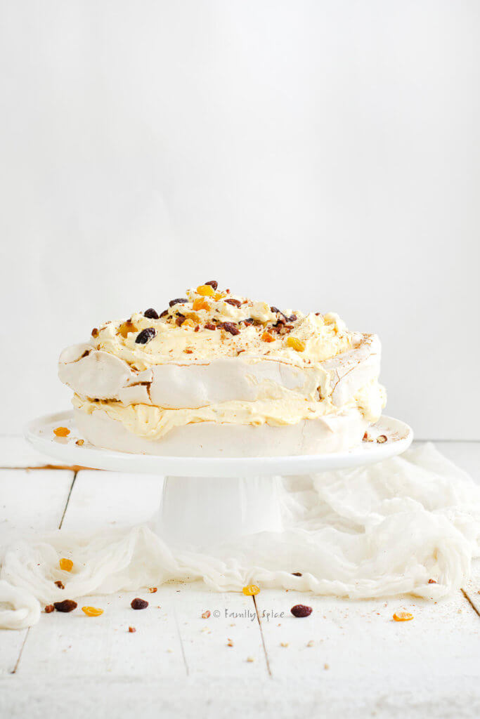 Side view of a pavlova cake with pumpkin whipped cream between the two layers and on top, garnished with chopped pecans and red and gold raisins