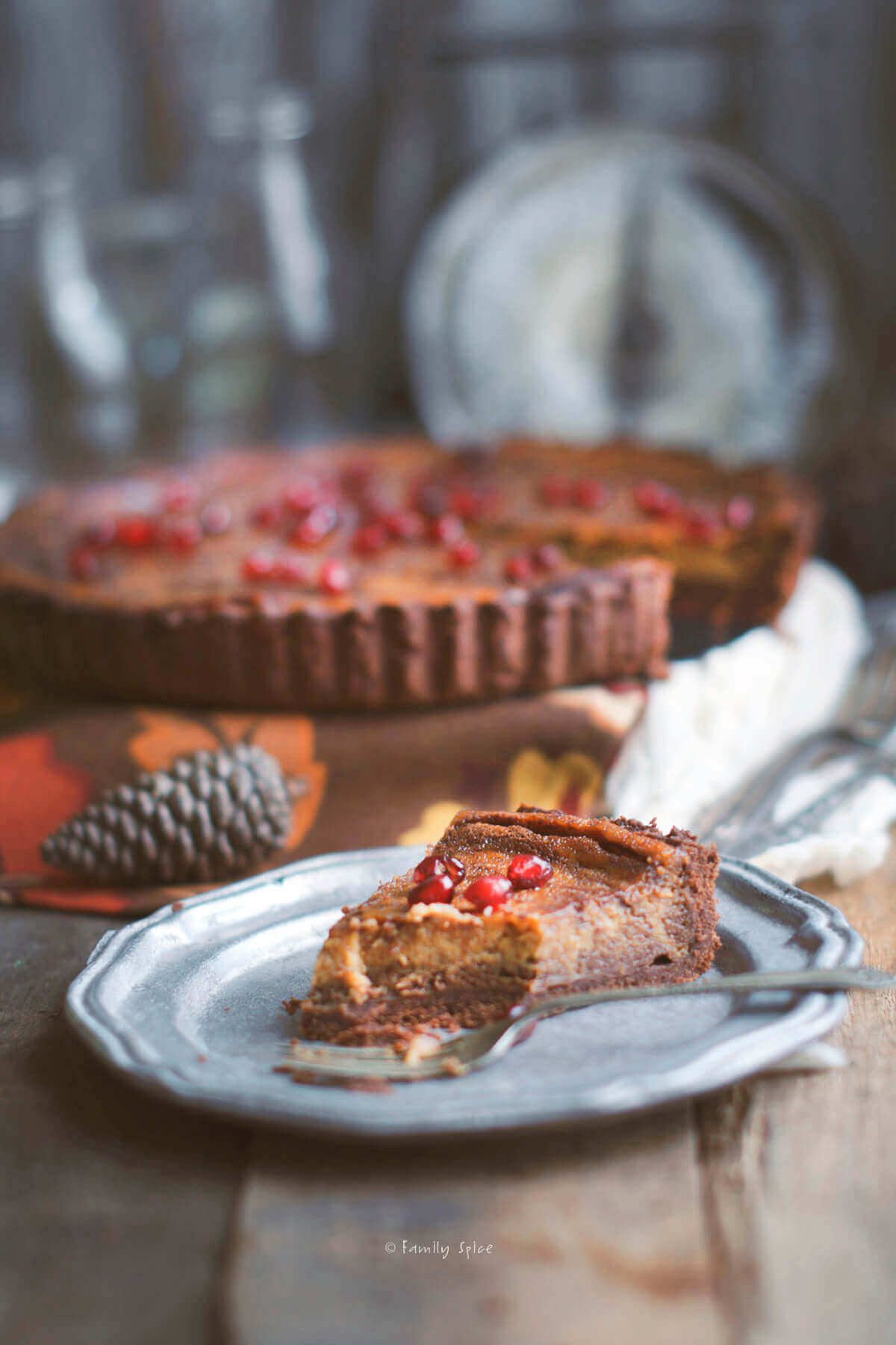 A slice of of a pumpkin pie with chocolate swirls baked in a chocolate pie shell topped with pomegranate arils with the rest of the pie in the background