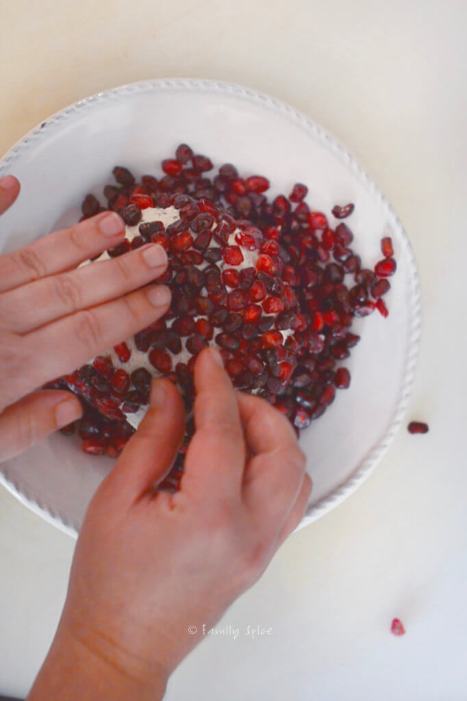 Hands pressing individual pomegranate arils into cheese ball on a white plate