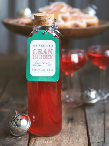 Closeup of a bottle of cranberry liqueur with two small stemmed glasses filled with it and cookies behind it
