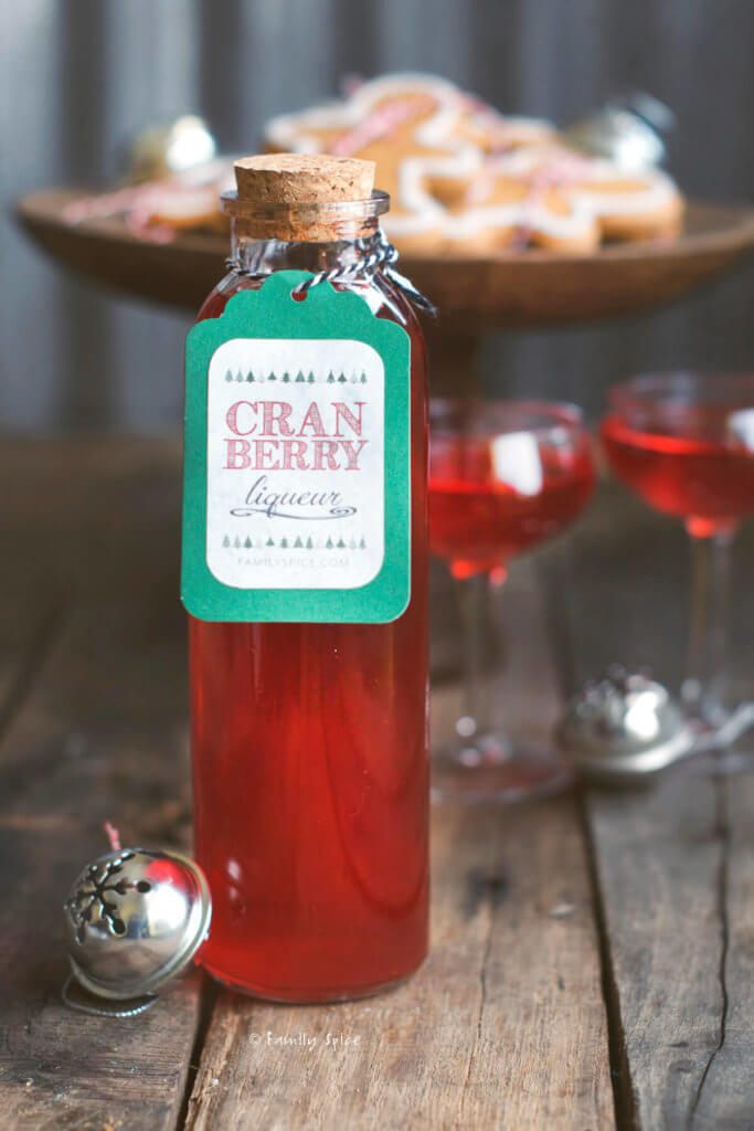 Closeup of a bottle of cranberry liqueur with two small stemmed glasses filled with it and cookies behind it