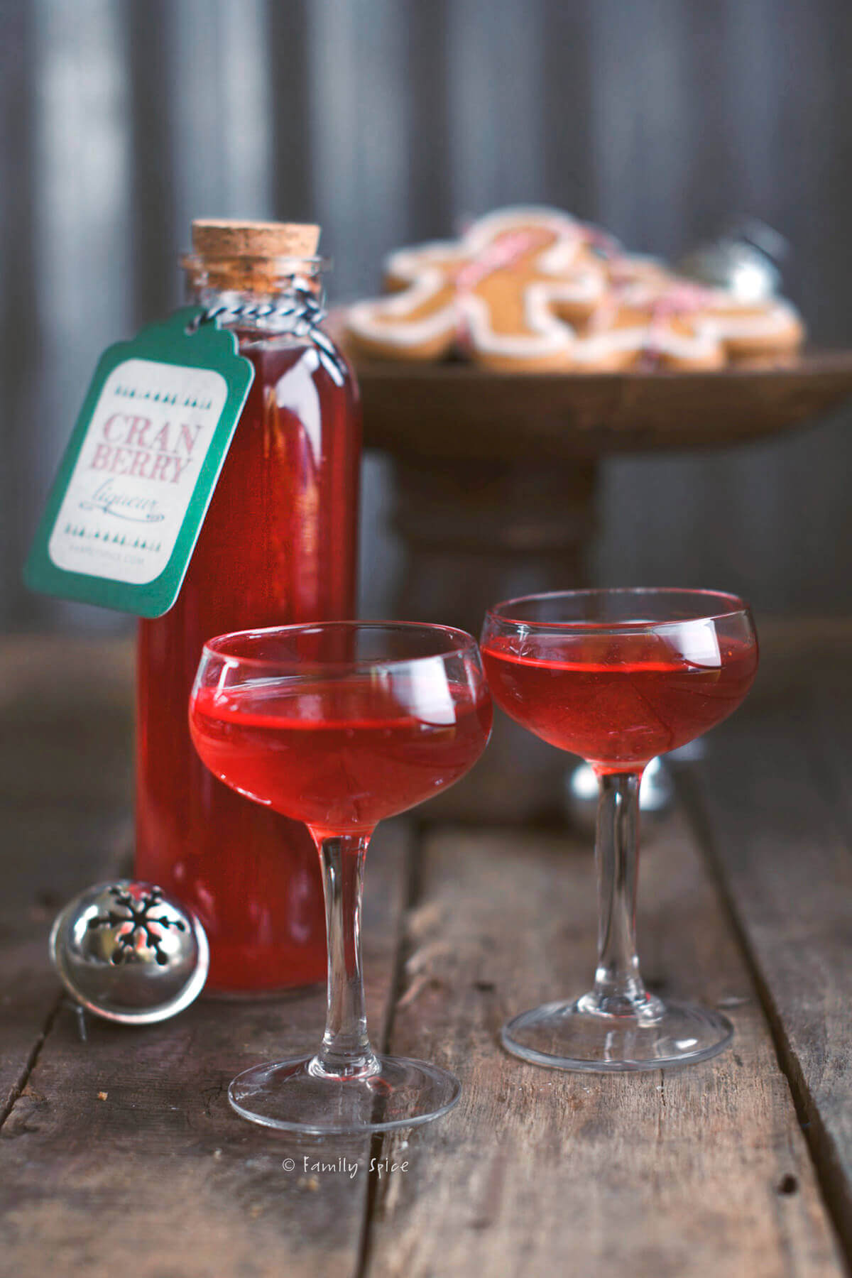 A bottle of cranberry liqueur with two small stemmed glasses filled with it and cookies behind it
