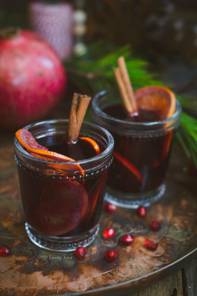 Closeup of two glasses of pomegranate mulled wine garnished with orange slices and cinnamon sticks on a dark rustic background