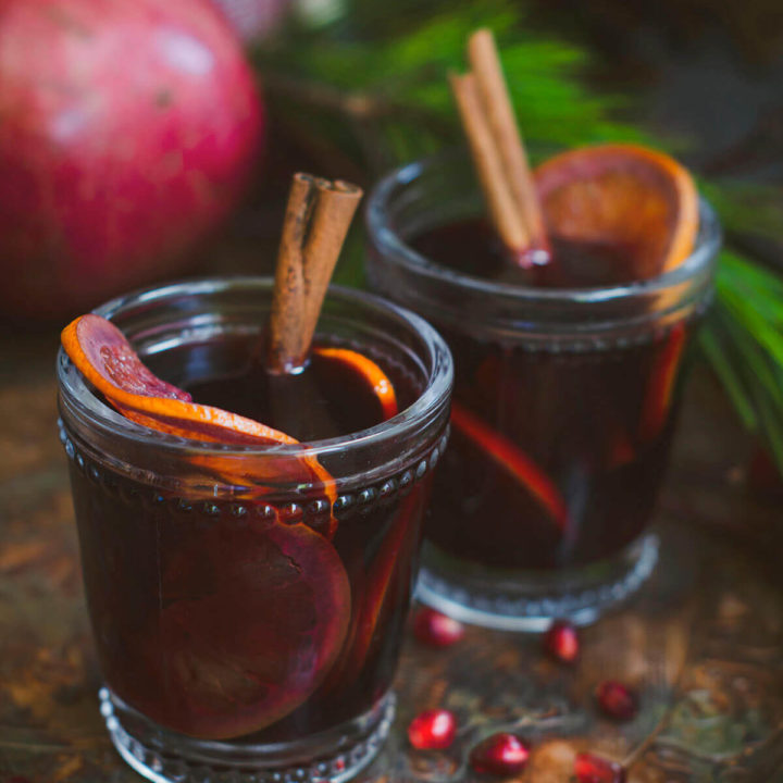 Closeup of two glasses of pomegranate mulled wine garnished with orange slices and cinnamon sticks on a dark rustic background