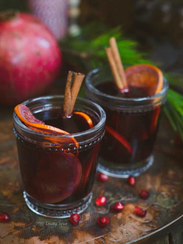 Closeup of two glasses of pomegranate mulled wine garnished with orange slices and cinnamon sticks on a dark rustic background