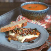Toasted brie sandwich with caramelized onions and raisins on a gray plate with a bowl of tomato soup behind it