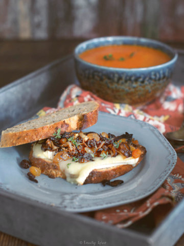 Toasted brie sandwich with caramelized onions and raisins on a gray plate with a bowl of tomato soup behind it