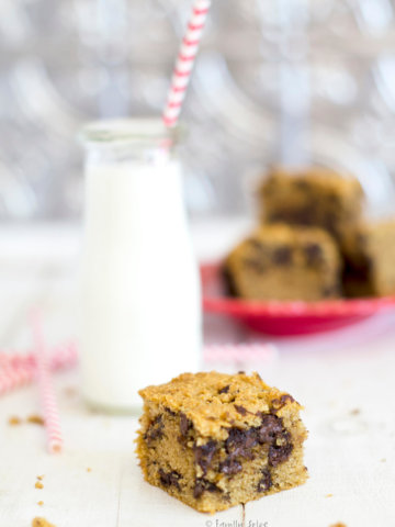 Closeup of a chocolate chip cookie bar with a plate full of them and a bottle of milk behind it