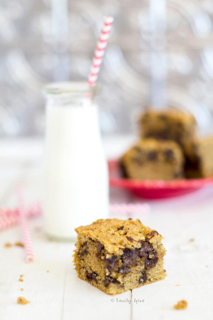 Closeup of a chocolate chip cookie bar with a plate full of them and a bottle of milk behind it