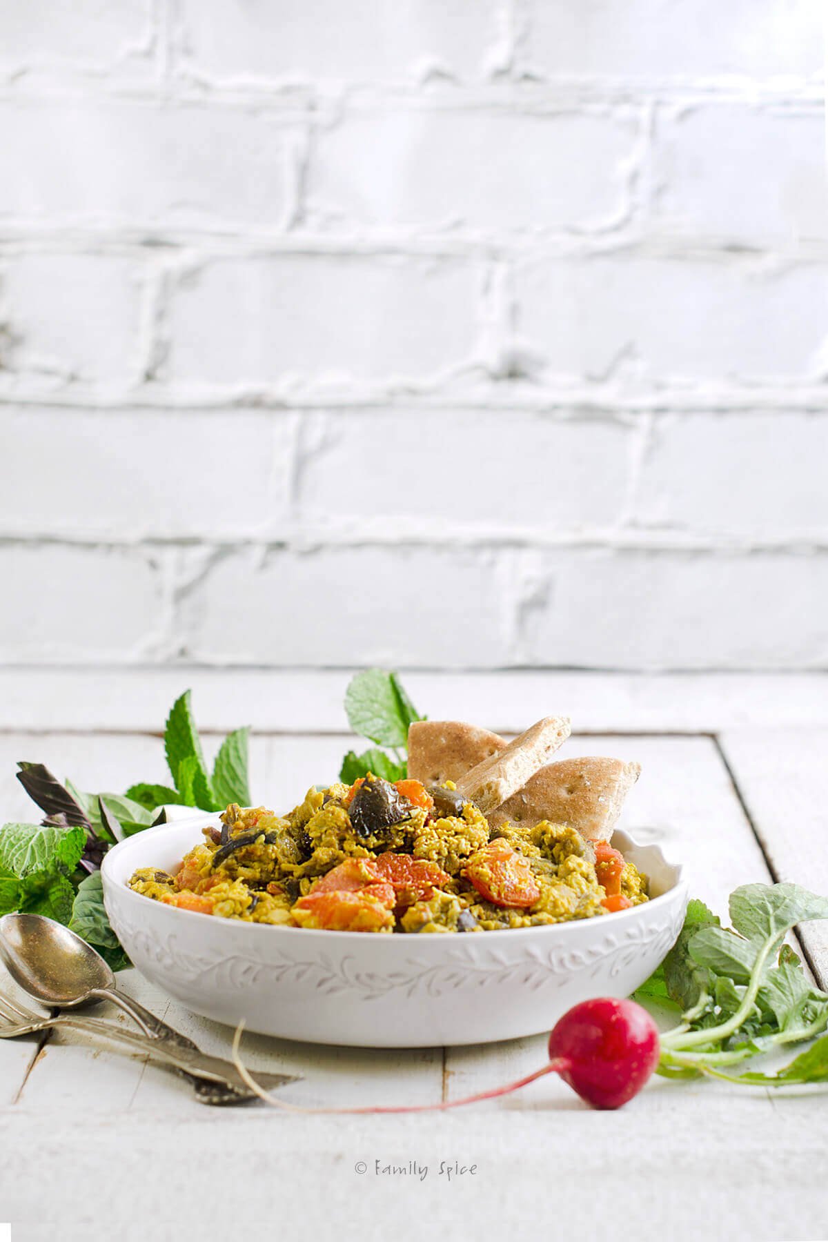 Side view of a white bowl with Mirza Ghasemi in it (a Persian egg dish) and herbs next to it