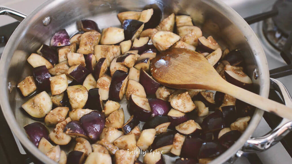 A stainless steel pan on the stove with chopped eggplant being browned in it