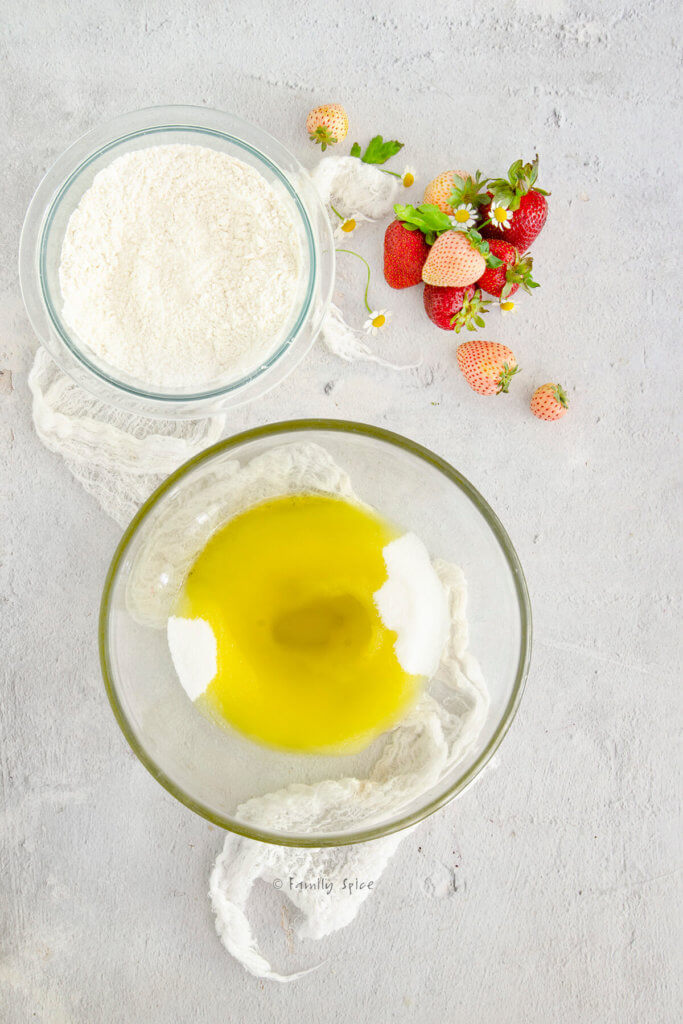A glass mixing bowl with olive oil and sugar in it and another bowl with flour mixture in it