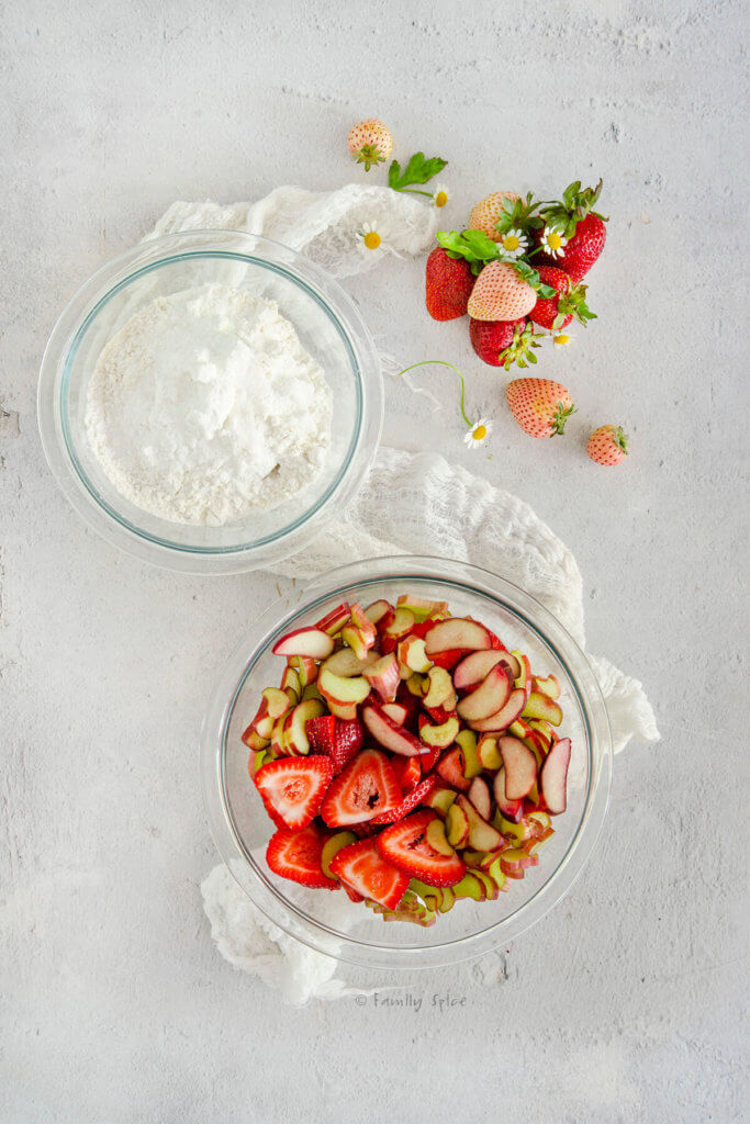 A bowl of chopped strawberries and rhubarb and another bowl with flour and dry ingredients in it