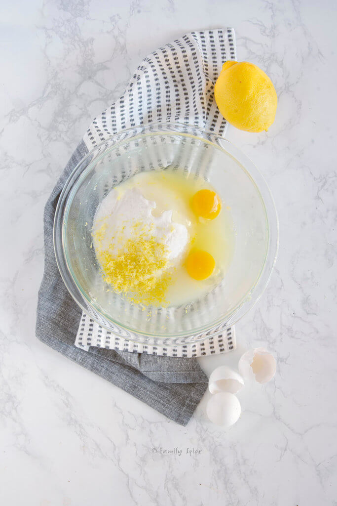 Eggs, sugar, lemon juice and zest in a glass mixing bowl with a lemon next to it