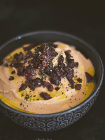 Closeup of a dark charcoal bowl with pumpkin hummus in it and garnished with olives and olive oil