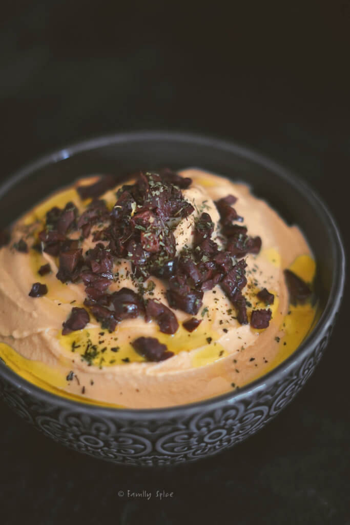 Closeup of a dark charcoal bowl with pumpkin hummus in it and garnished with olives and olive oil