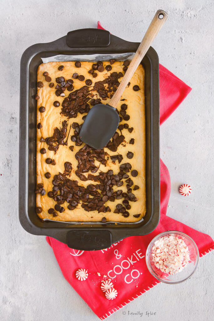 Shortbread cookie batter freshly baked in a 9x13 metal pan with chocolate chips melting over it and spread with rubber spatula
