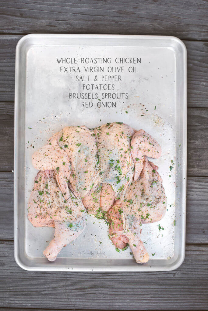 Top view of a raw spatchcocked chicken smothered in herbs on a baking sheet with ingredients needed for recipe labeled
