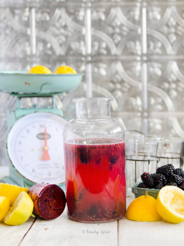 Side view of a bottle filled with blackberry lemonade with lemons, blackberries and an antique scale around it