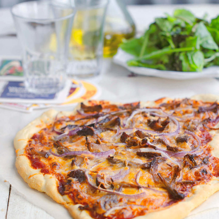 A rustic pulled pork pizza with olive oil and arugula behind it