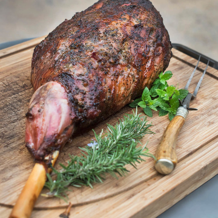 Side view of a grilled leg of lamb on a wood cutting board with fresh herbs next to it