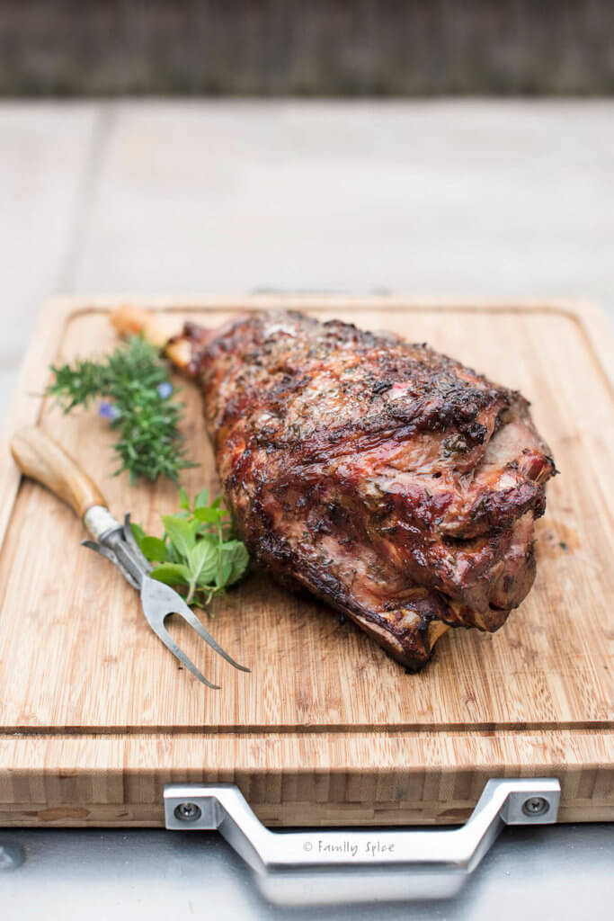 Top of a grilled leg of lamb on a wood cutting board with fresh herbs and serving fork next to it