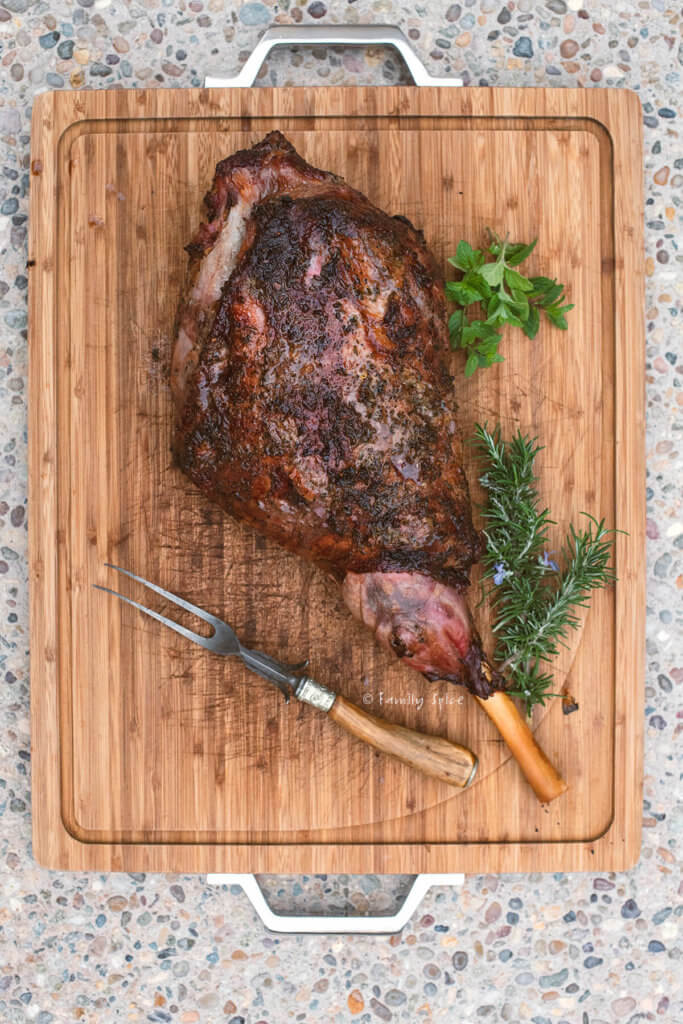 Top view of a grilled leg of lamb on a wood cutting board with fresh herbs next to it