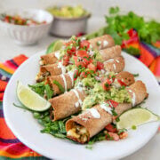Closeup of baked steak taquitos on a white plate garnished with guacamole, pico de gallo and sour cream.