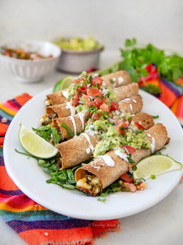 Closeup of baked steak taquitos on a white plate garnished with guacamole, pico de gallo and sour cream.