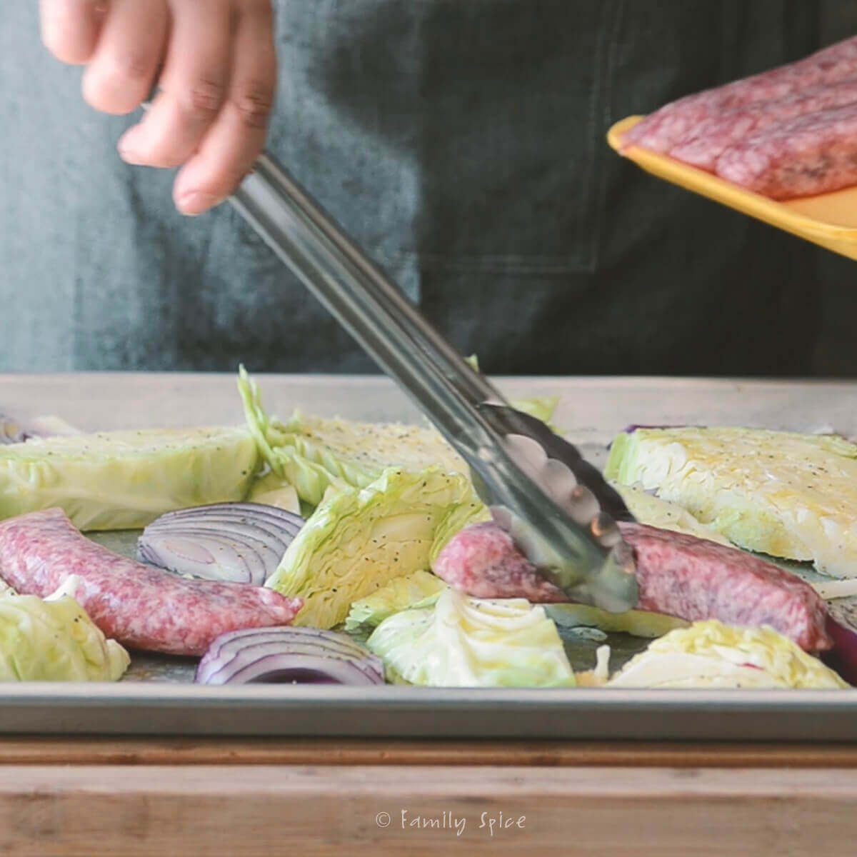 Using tongs to place brats on a baking sheet with wedges of cabbage and red onions